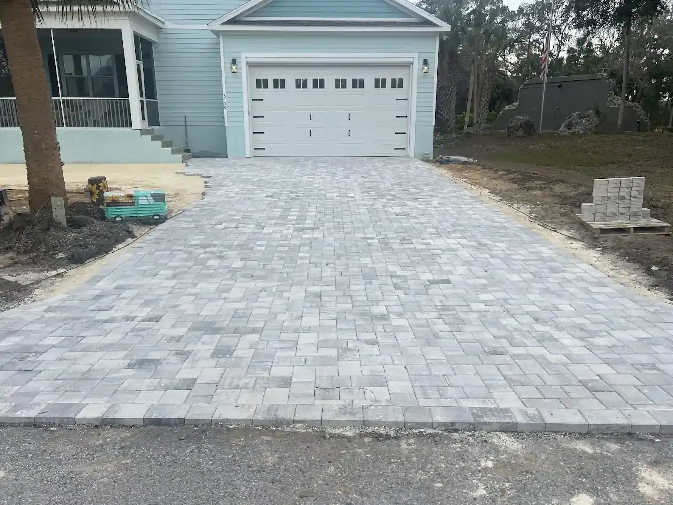 A newly installed gray brick driveway leads to a two-car garage of a light blue house in Hernando County; construction materials and unfinished edges are visible on both sides of the driveway.