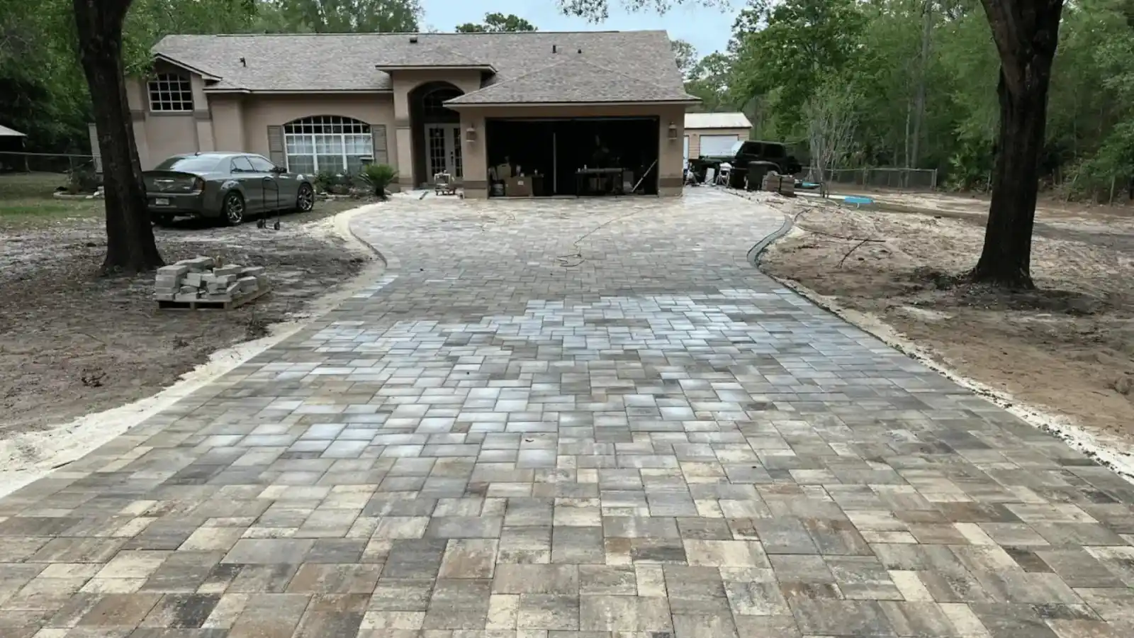 Newly installed paver driveway leading to a beige house with an open garage in Sumter, FL; cars are parked on both sides, and the surrounding yard appears unfinished with exposed soil.