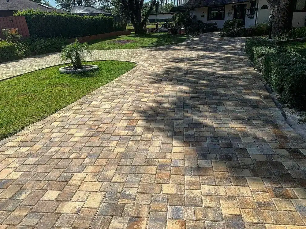 A paved driveway made of multi-colored bricks curves through a green lawn toward a house, with a small circular garden by a Landscaper Citrus expert on the left side. Trees cast shadows across the driveway in sunny FL.