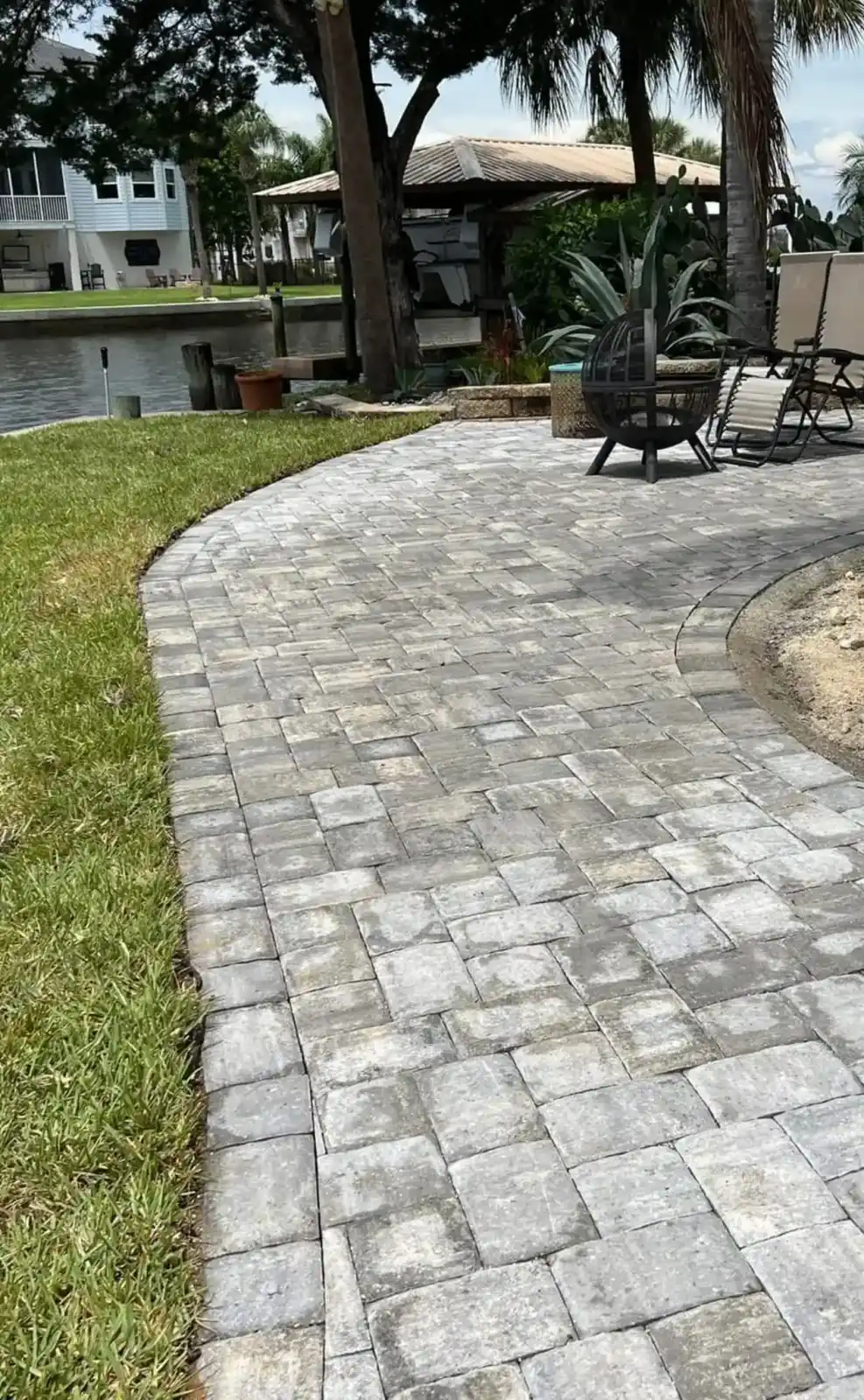 A curved stone patio walkway bordered by grass leads to outdoor chairs and a fire pit, designed by a skilled Hernando County landscaper. A canal, trees, and houses are visible in the background, creating a relaxing FL retreat.