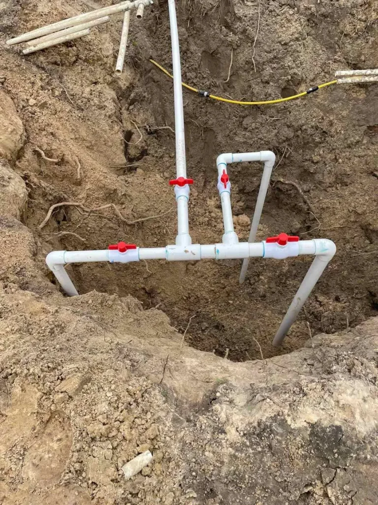 A section of white PVC plumbing pipes with red-handled valves is installed underground in a dirt trench in Hernando County, surrounded by soil and exposed roots.