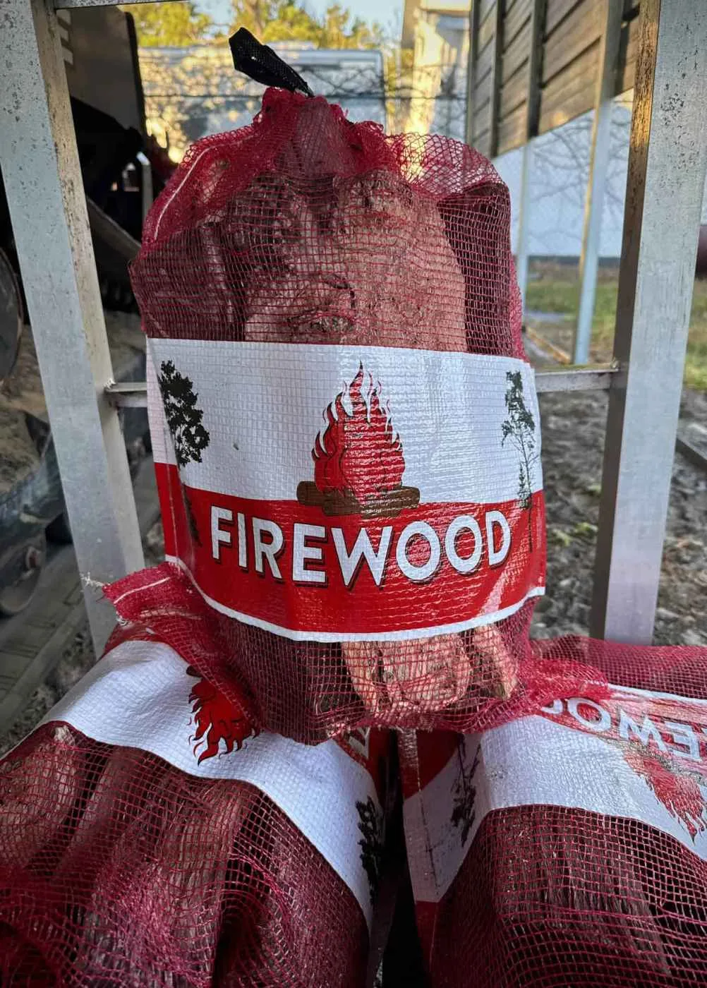 A red mesh bag filled with firewood sits atop similar bags outdoors near a fence in Hernando County, FL. The bag features a white label with a red flame and bold "FIREWOOD" print—ideal for any landscaper in Citrus or surrounding areas.