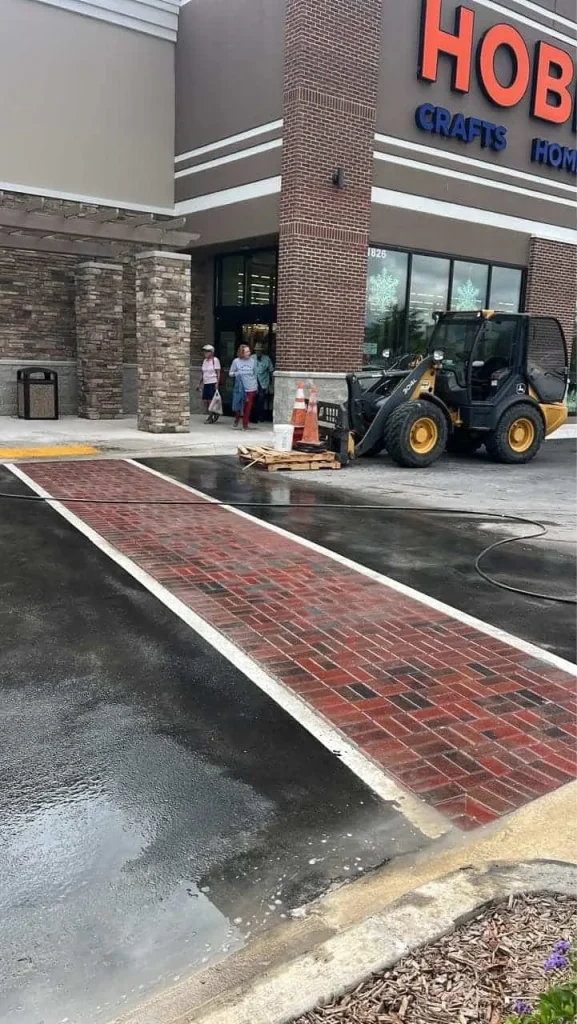 A skid steer with a pallet and orange cone is parked in front of a Hobby Lobby store entrance in Hernando County, near a red brick crosswalk on wet pavement. Several people stand by the entrance.