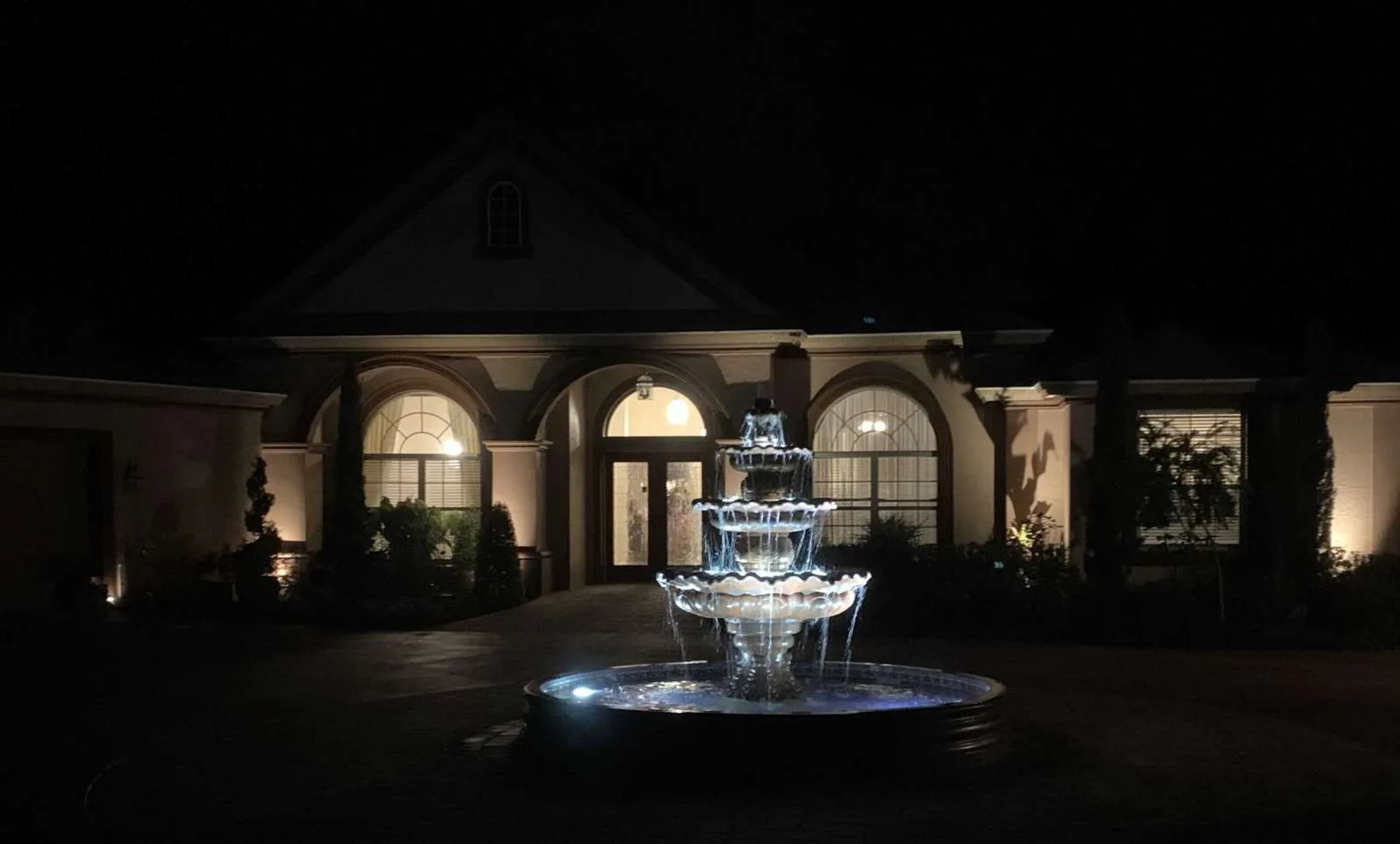 A three-tiered fountain is illuminated at night in front of a large Hernando County, FL home with arched windows and a lit entryway, creating a dramatic and elegant scene.