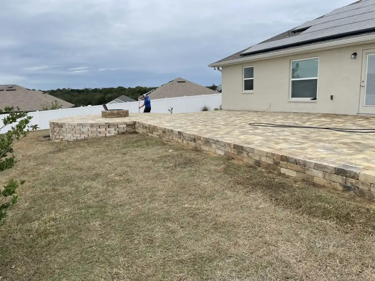 A backyard in Hernando County, FL, with a newly built stone patio and low retaining wall by a beige house; a person stands near the wall, and a garden hose lies on the patio. The lawn is patchy under cloudy skies—work by Landscaper Citrus.