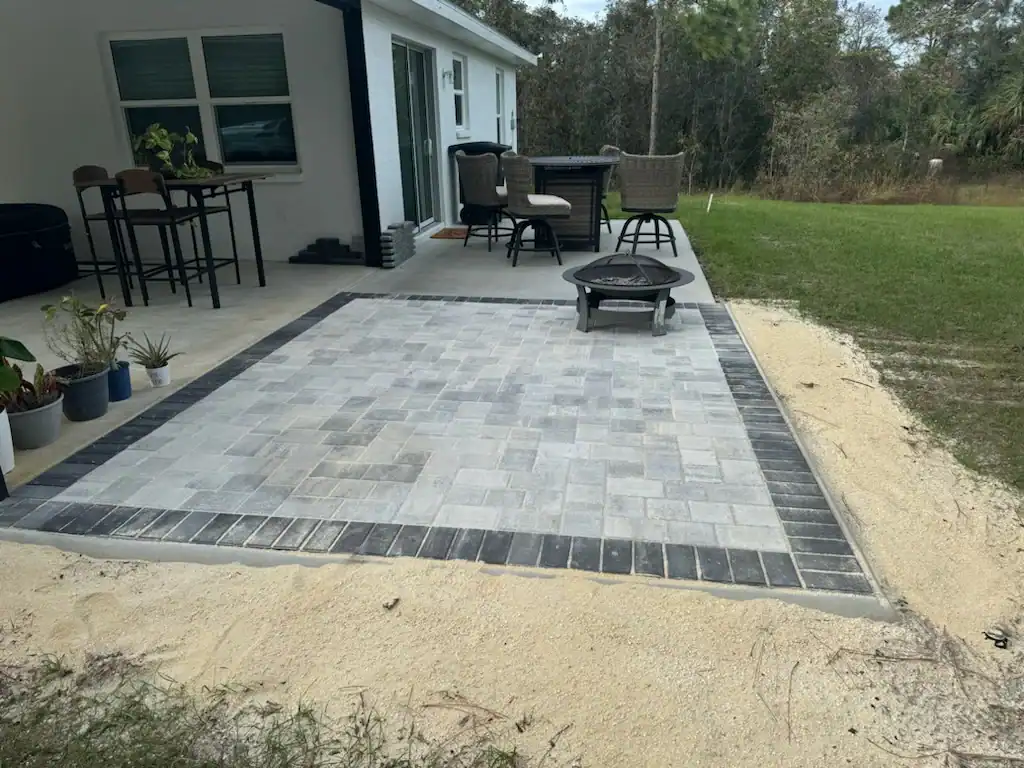 A backyard patio in Hernando County with gray and dark border pavers, surrounded by sand, features potted plants, a tall table with chairs, a fire pit, and a grassy yard next to a house with sliding glass doors.