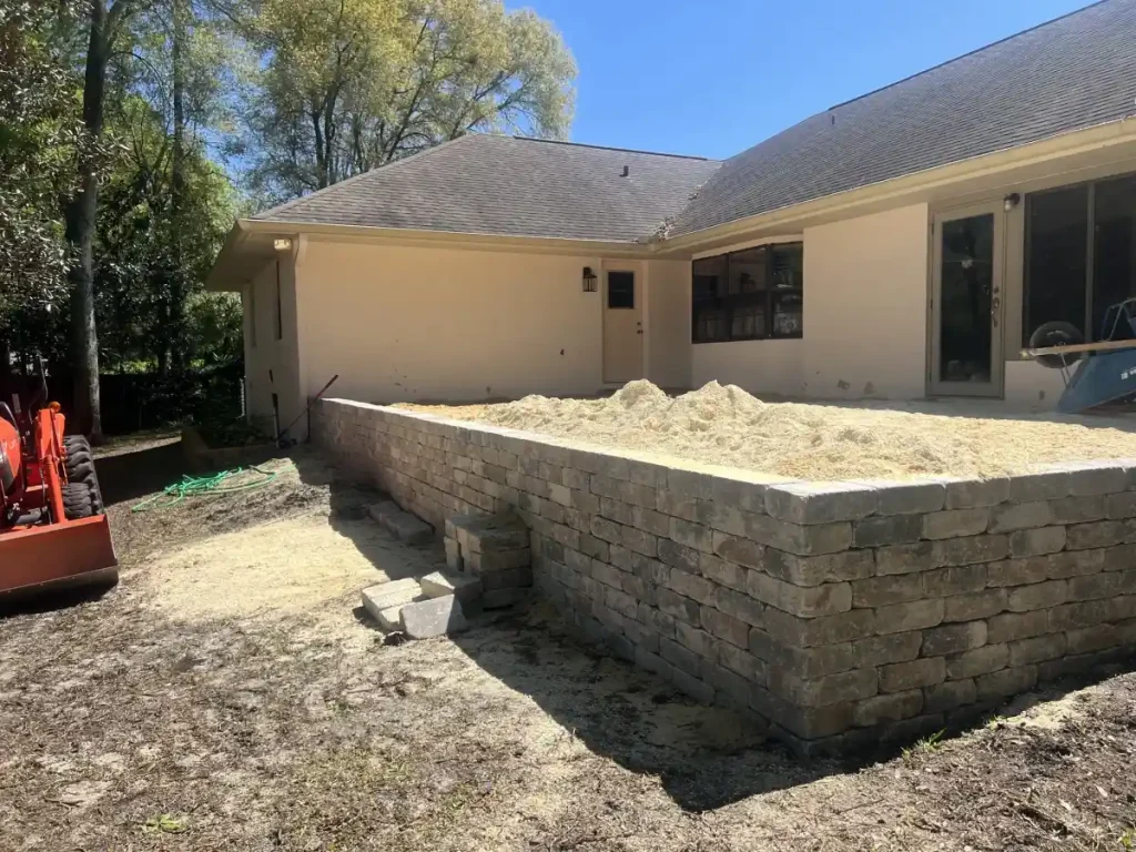 A raised stone retaining wall filled with sand sits beside a light-colored house. A few stone steps lead up to the area, with construction equipment nearby—typical work by an expert landscaper in Citrus, FL or Sumter.