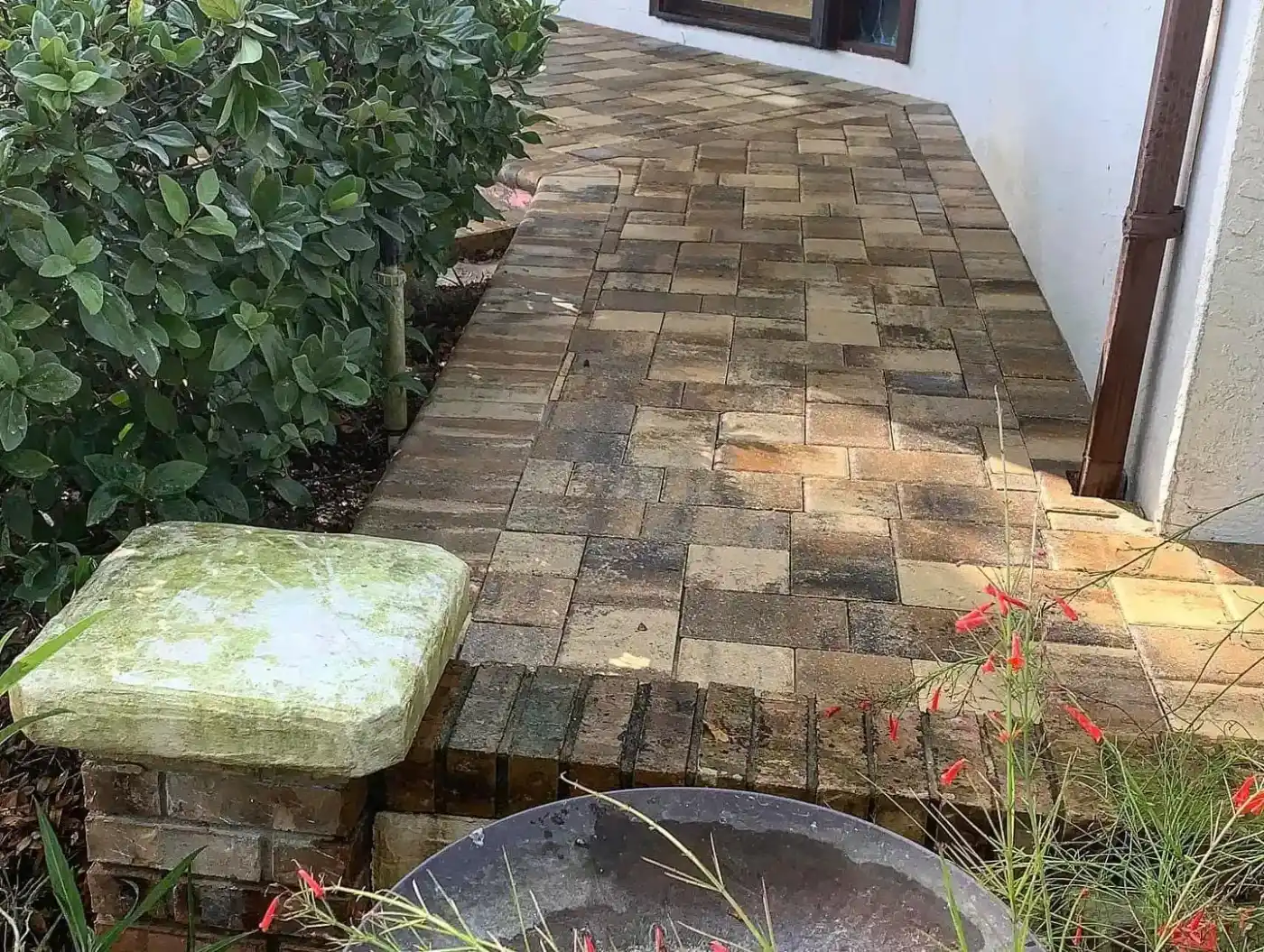 A narrow walkway made of interlocking stone pavers runs alongside a white building, bordered by green shrubs and a small garden with red flowers—expertly designed by a top landscaper in Hernando County, FL.