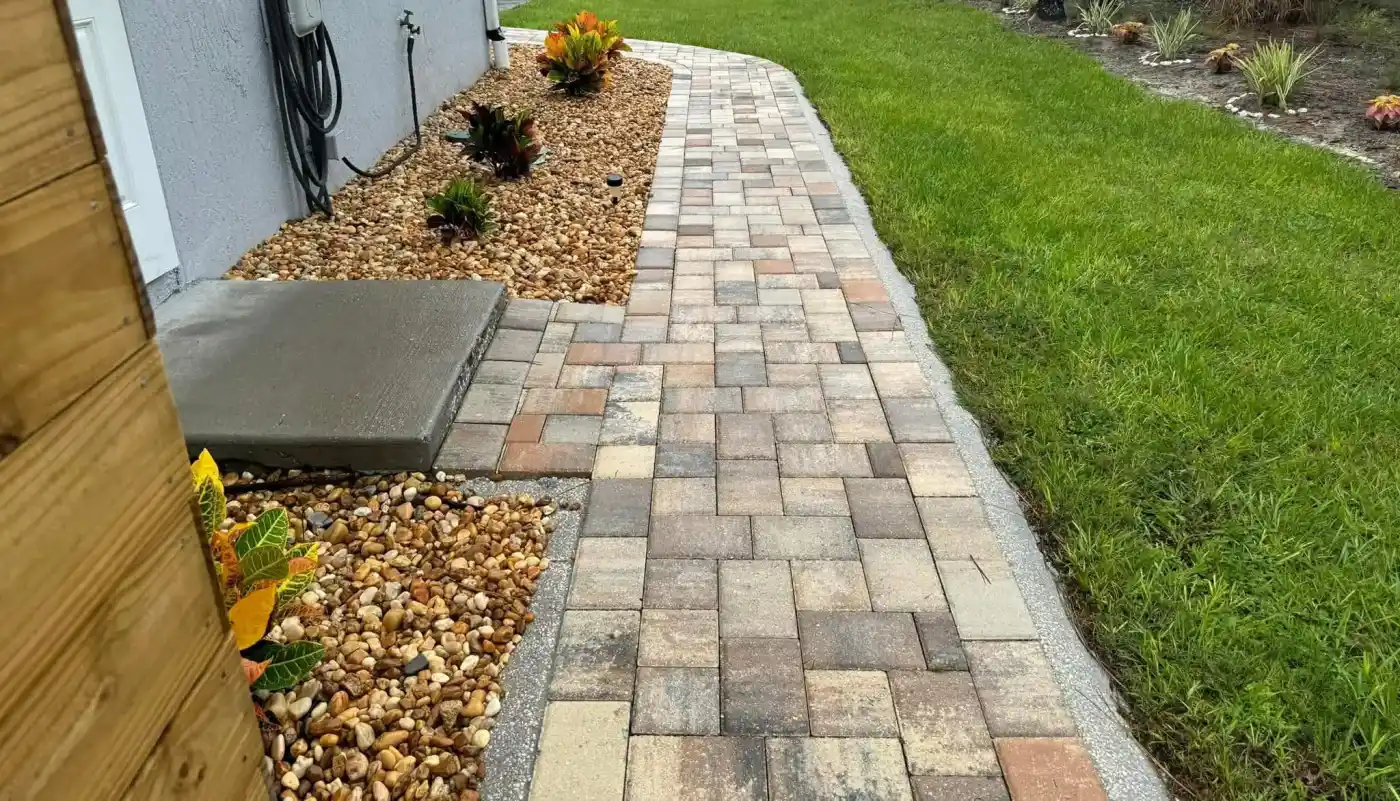 A paved brick walkway bordered by landscaping stones and small colorful plants runs alongside a gray house in Hernando County, FL, with a green lawn on the right.