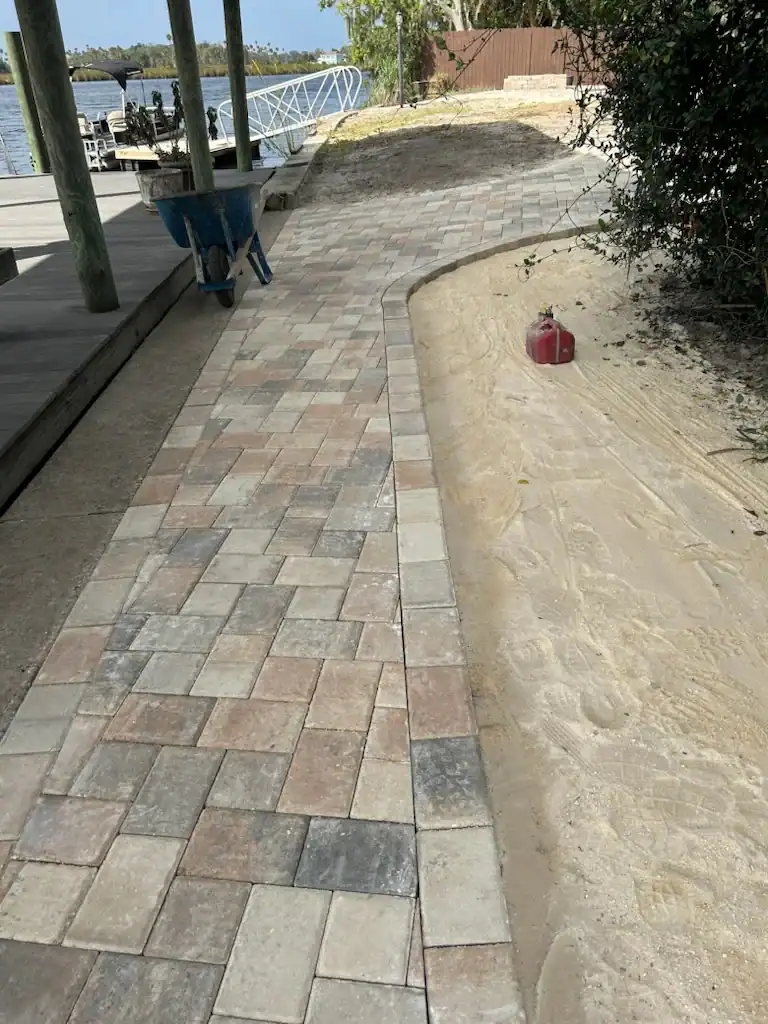A paved walkway made of rectangular stone tiles runs alongside a sandy area with a red gas can in Hernando County, FL. A blue wheelbarrow rests on the path, and a dock extends over the water in the background.