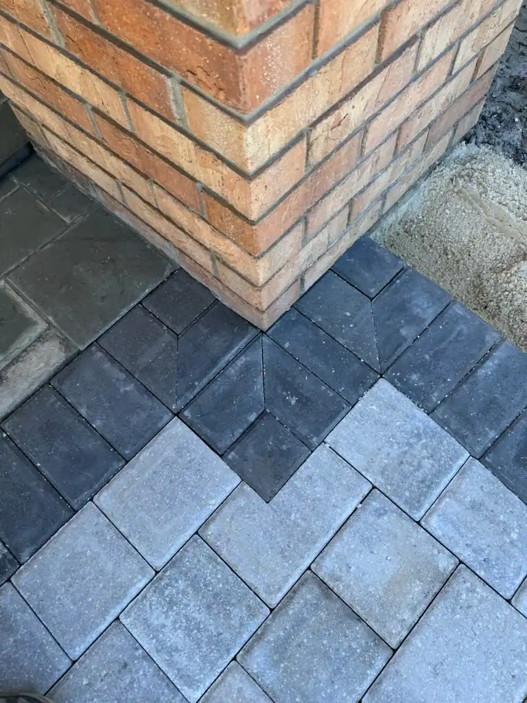 Gray paving stones are precisely arranged in a chevron pattern meeting at the corner of a tan brick wall, with sand visible in the upper right corner—expertly installed by Landscaper Citrus in Hernando County.