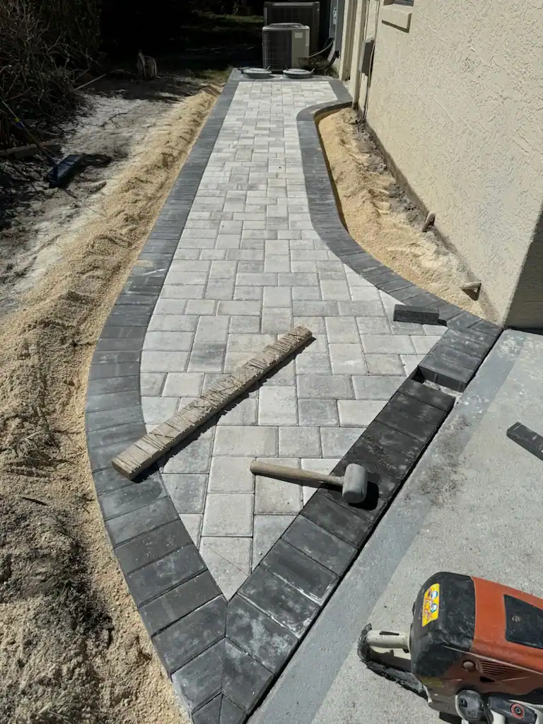 A partially completed walkway made of light gray rectangular pavers with a dark gray border, constructed by a skilled Hernando County landscaper, with construction tools and sand along the edges.