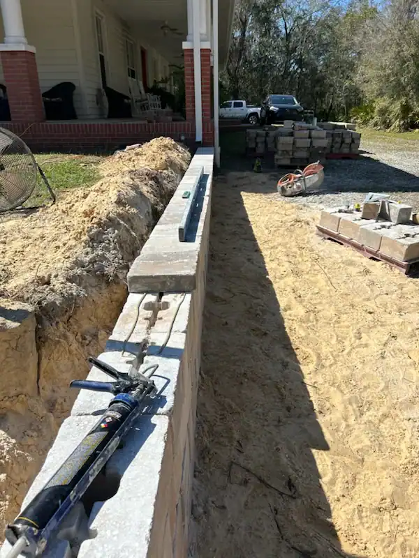 A partially constructed retaining wall runs parallel to a house with a porch in Sumter, FL. Tools and construction materials, including bricks and a caulking gun, are on and around the wall, ready for Landscaper Citrus to continue work.
