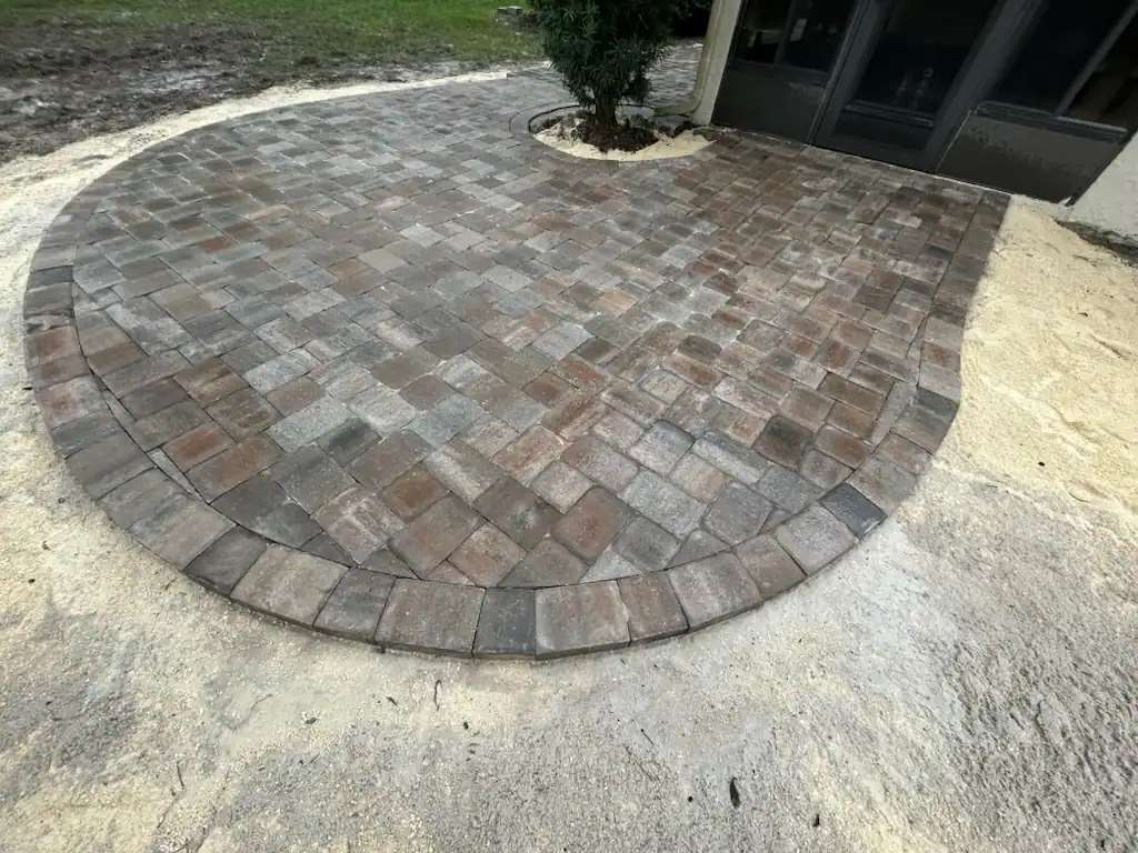 A circular patio made of interlocking stone pavers with a curved edge, adjacent to a building, features a small tree planted in a cutout. The surrounding area shows unfinished landscaping with exposed sand.