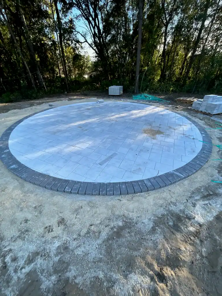 A circular patio made of light-colored pavers with a dark border is set in a sandy area, surrounded by trees. Stacks of unused pavers and construction materials are visible nearby, showcasing the work of a skilled Hernando County, FL landscaper.