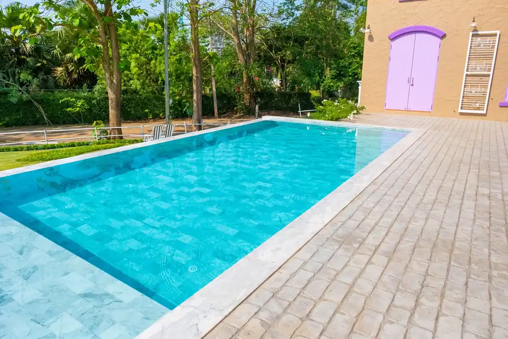 A rectangular outdoor swimming pool with clear blue water, surrounded by light-colored stone tiles designed by a top landscaper in Citrus. Trees and greenery are in the background, and a building with a purple door is on the right.