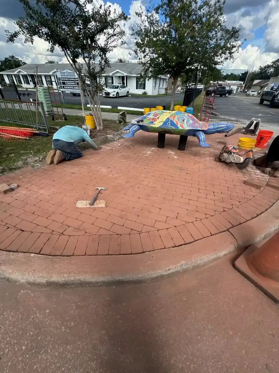 A Landscaper in Citrus County kneels on a newly paved brick area next to a colorful turtle sculpture, working with bricks and tools. Residential buildings and parked cars are visible in the background.