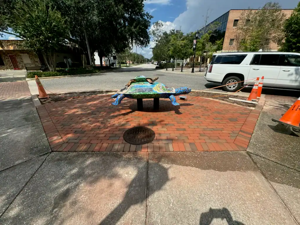 A colorful turtle sculpture is displayed on a brick platform at a street corner in Sumter, FL, surrounded by orange traffic cones. Trees, a white SUV, and buildings are visible in the background on a partly cloudy day.