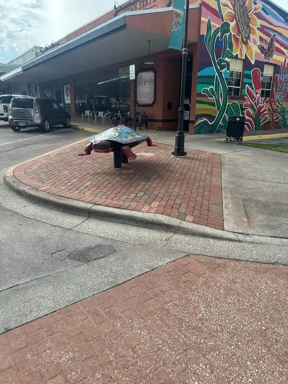A turtle sculpture is displayed on a brick sidewalk near a colorful mural with flowers on a building wall in Citrus, FL. Several parked cars, adding to the scene’s charm in Hernando County, are visible in the background.