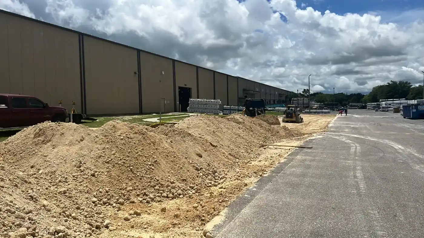 Large piles of dirt line a paved road beside a long beige warehouse under a partly cloudy sky in Hernando County, with construction equipment and materials from Landscaper Citrus visible in the background.