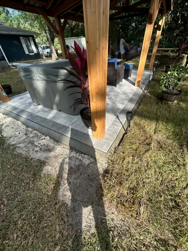 A covered patio with wooden posts, a hot tub, potted plants, and outdoor furniture on a tiled platform. Sunlight casts shadows on the grass and patio area, landscaped by experts serving Citrus, Sumter, and Hernando County. Trees and a house in the background.