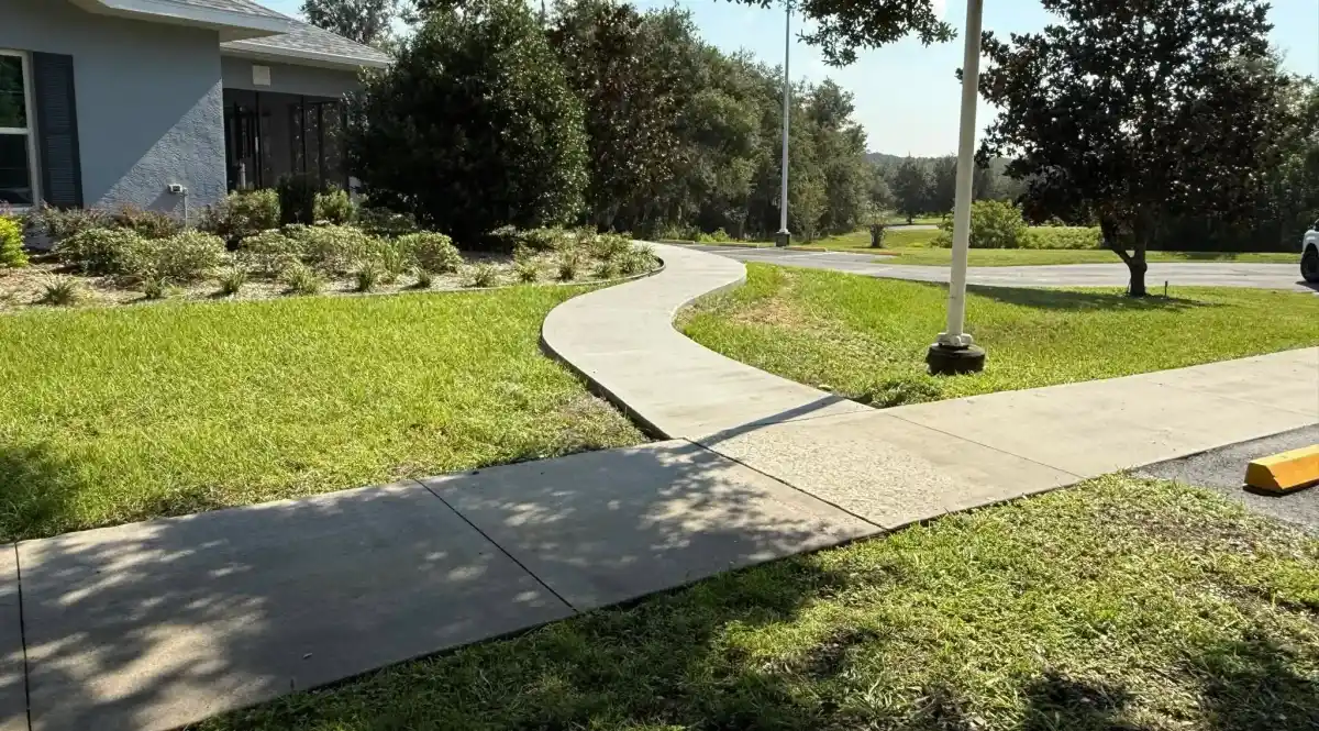 A curved concrete sidewalk winds through a neatly maintained grassy yard in Sumter, FL, beside a house bordered by hedges and trees, with a paved driveway visible—showcasing the expert touch of Landscaper Citrus.