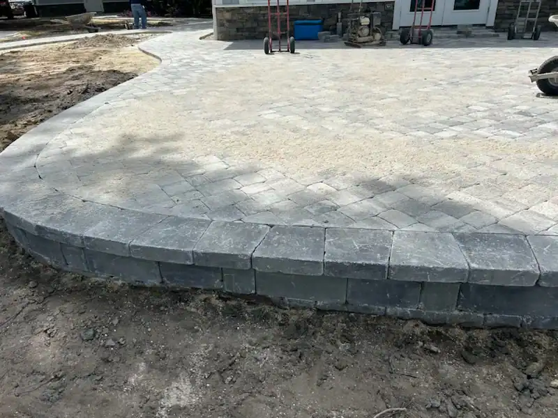 A newly constructed stone paver patio with a raised edge and unfinished surrounding dirt, built by a skilled landscaper in Citrus, FL, sits in front of a building under construction in Hernando County.