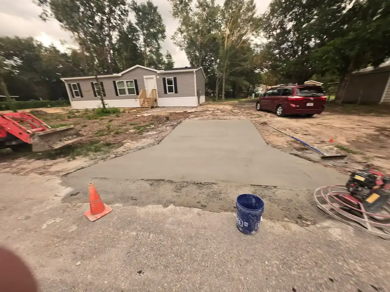 Freshly poured concrete driveway in Hernando County leads to a single-story house with a porch. An orange traffic cone, blue bucket, and construction equipment sit out front, with a red car parked on the right.