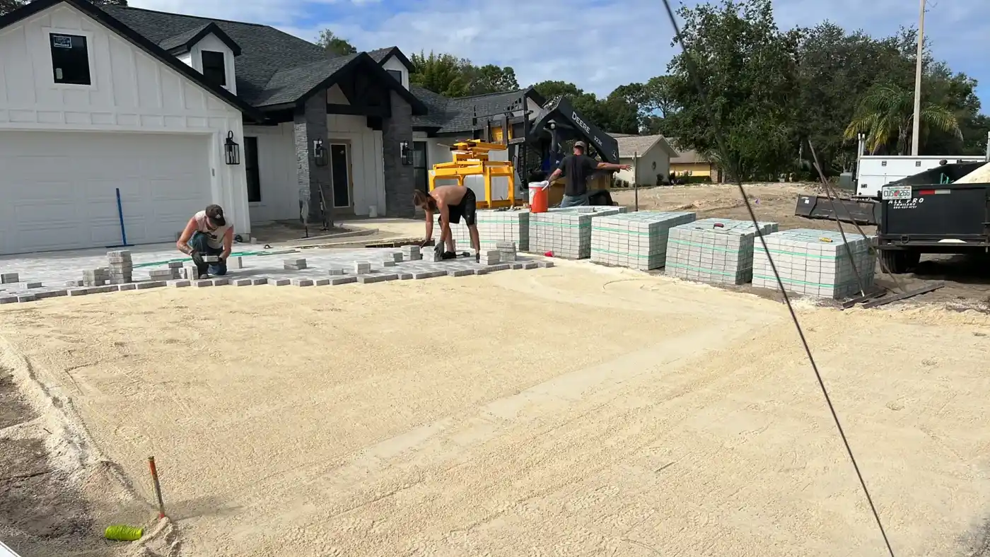 Workers lay pavers for a driveway in front of a modern white house under construction in Hernando County, FL. Piles of pavers, tools, and equipment are visible around the site on a clear day.