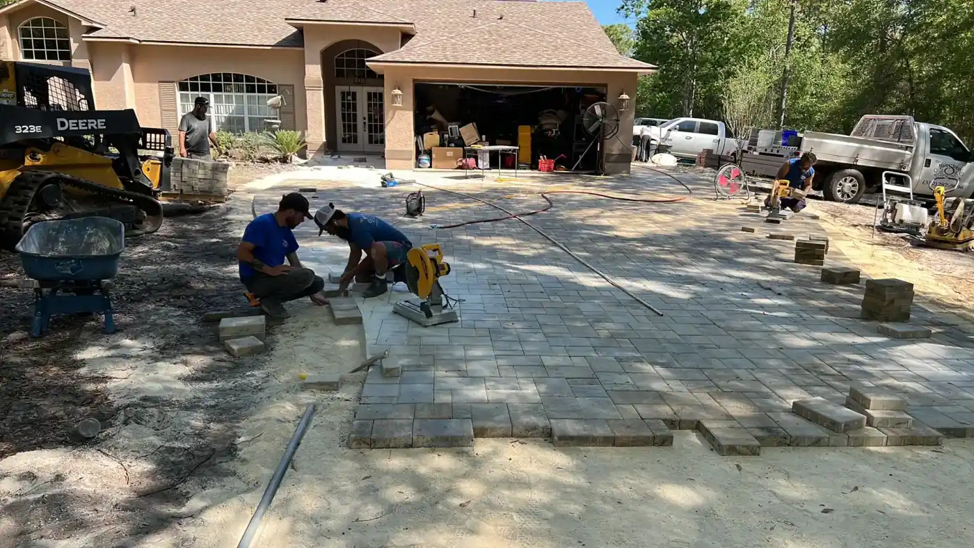 Two workers from a Landscaper Citrus team are installing pavers on a driveway in Hernando County, FL. Construction equipment and tools are nearby, and the garage door is open with more equipment visible inside. The driveway is partially completed.