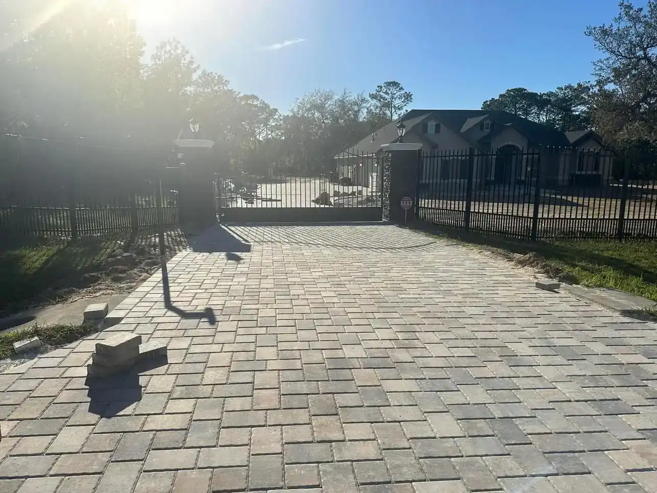 A gated driveway with a paved stone surface leads to a large house in Hernando County, FL, surrounded by a black metal fence. A few loose bricks are on the ground, while sunlight creates a bright glare in the upper left corner.