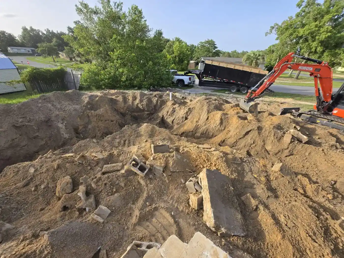 A large dirt hole with broken concrete pieces scattered inside, an orange excavator on the right, and a black dump trailer and white truck in the background—typical sights for a Landscaper Citrus job in sunny Sumter, FL, surrounded by trees and grass.
