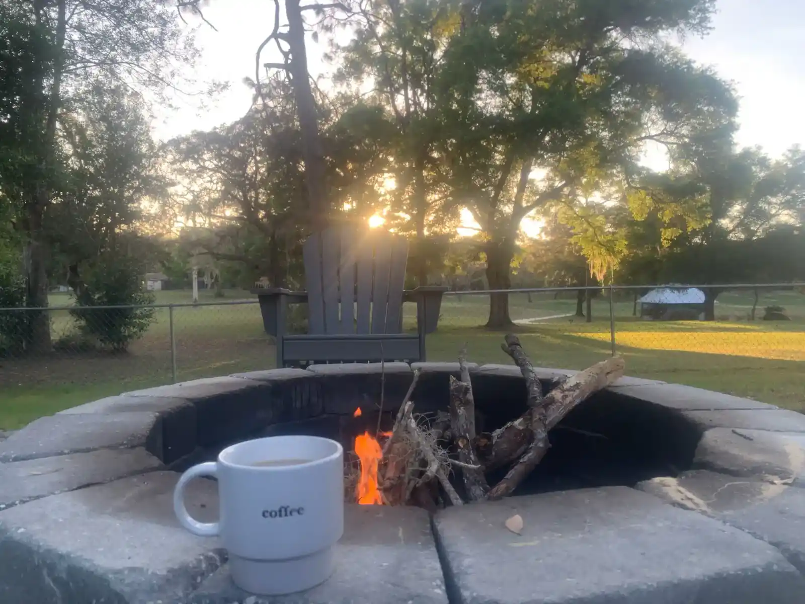 A white mug labeled "coffee" sits on the edge of a stone fire pit with a small fire burning inside. Behind the pit, an empty chair overlooks tall trees and grassy yard—typical of the tranquil FL landscapes in Sumter and Citrus counties at sunset.