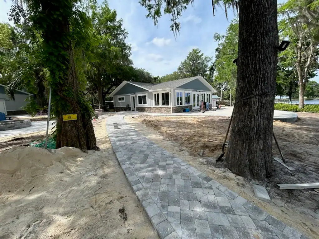 A gray house with large windows sits among trees, with a new stone walkway leading up to it. The sandy soil and scattered materials suggest ongoing landscaping work by a skilled landscaper in Citrus and Hernando County.