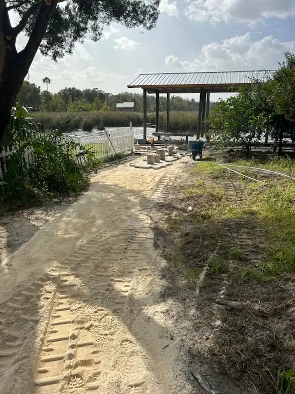 A sandy path with tire tracks leads to a dock by the water in Hernando County, FL. Pavers and construction materials are scattered nearby, with trees and a picket fence along the side under a partly cloudy sky.