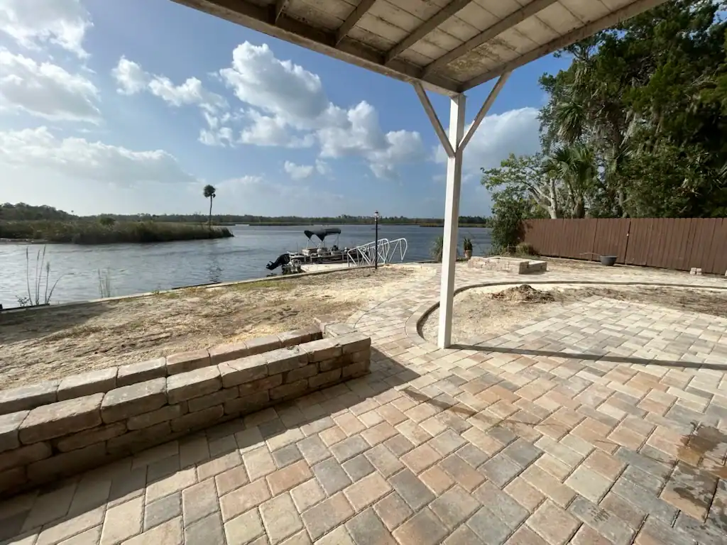 A paved patio with a partial roof overlooks a calm river in Hernando County, FL. A small boat is docked by the shore, and trees, grass, and a wooden fence create a scenic backdrop—perfect inspiration for any Landscaper Citrus project.