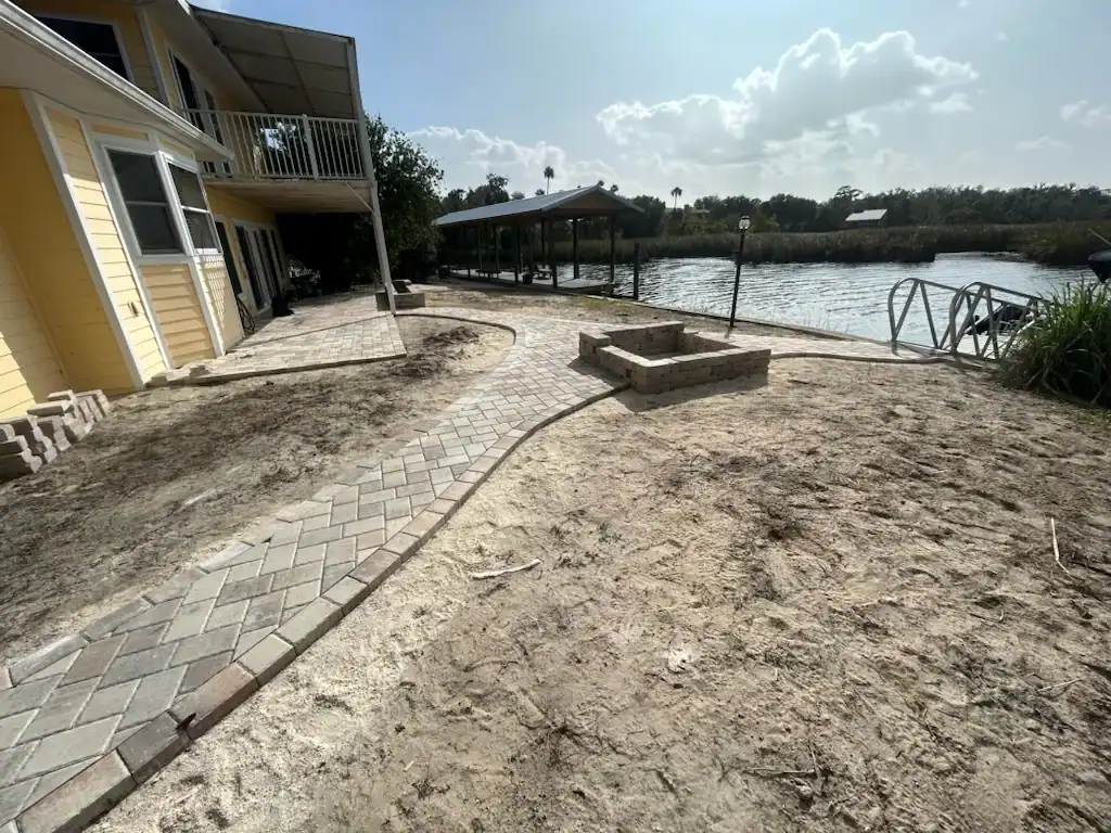 A backyard in Hernando County, FL, features a paved walkway leading to a square fire pit beside a yellow house. The sandy soil yard overlooks waterfront views, complete with a dock and covered boat area under a partly cloudy sky.