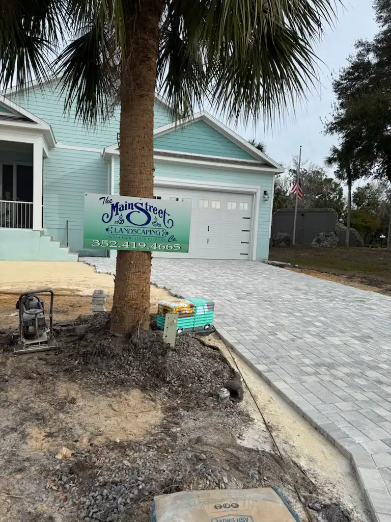 A freshly paved driveway leads to a light blue house with white trim in Hernando County, FL. A palm tree with a landscaping company sign stands at the edge, and landscaping materials are stacked nearby.
