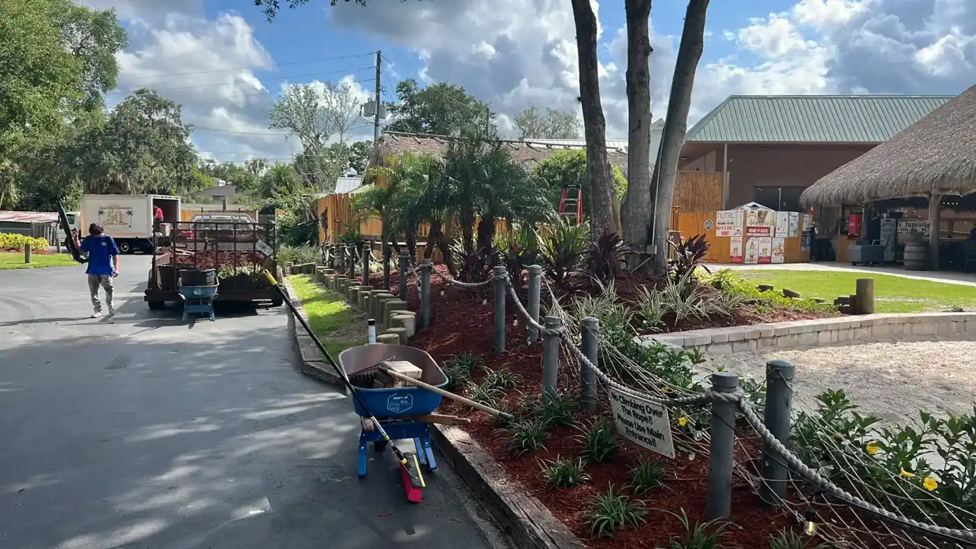 A landscaped garden with trees, plants, mulch, and a rope fence runs alongside a paved road in Hernando County, FL. A wheelbarrow and tools are in the foreground, while a person walks near trucks and buildings under a partly cloudy sky.