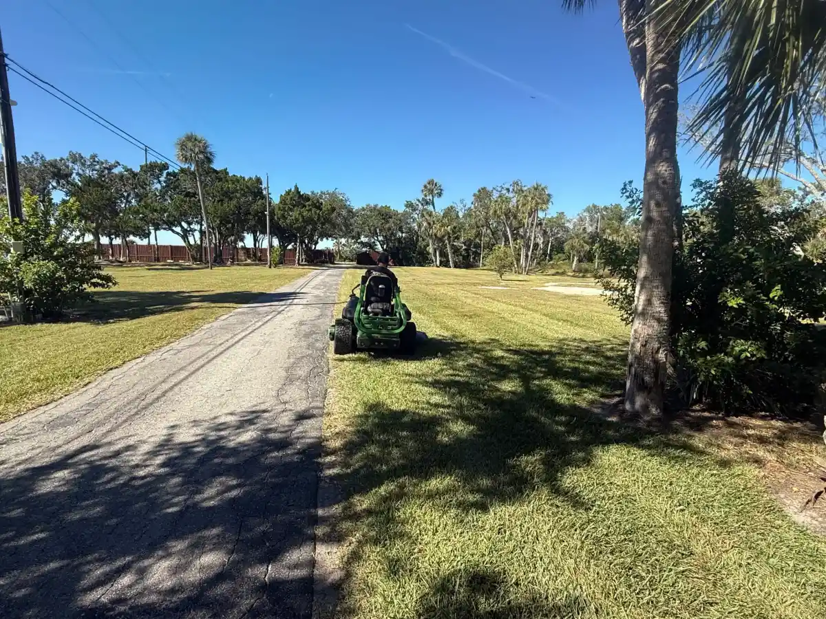 A landscaper rides a green lawn mower on grass beside a paved road under a clear blue sky, with trees and power lines in the background—typical of the scenic views in Sumter and Hernando County.