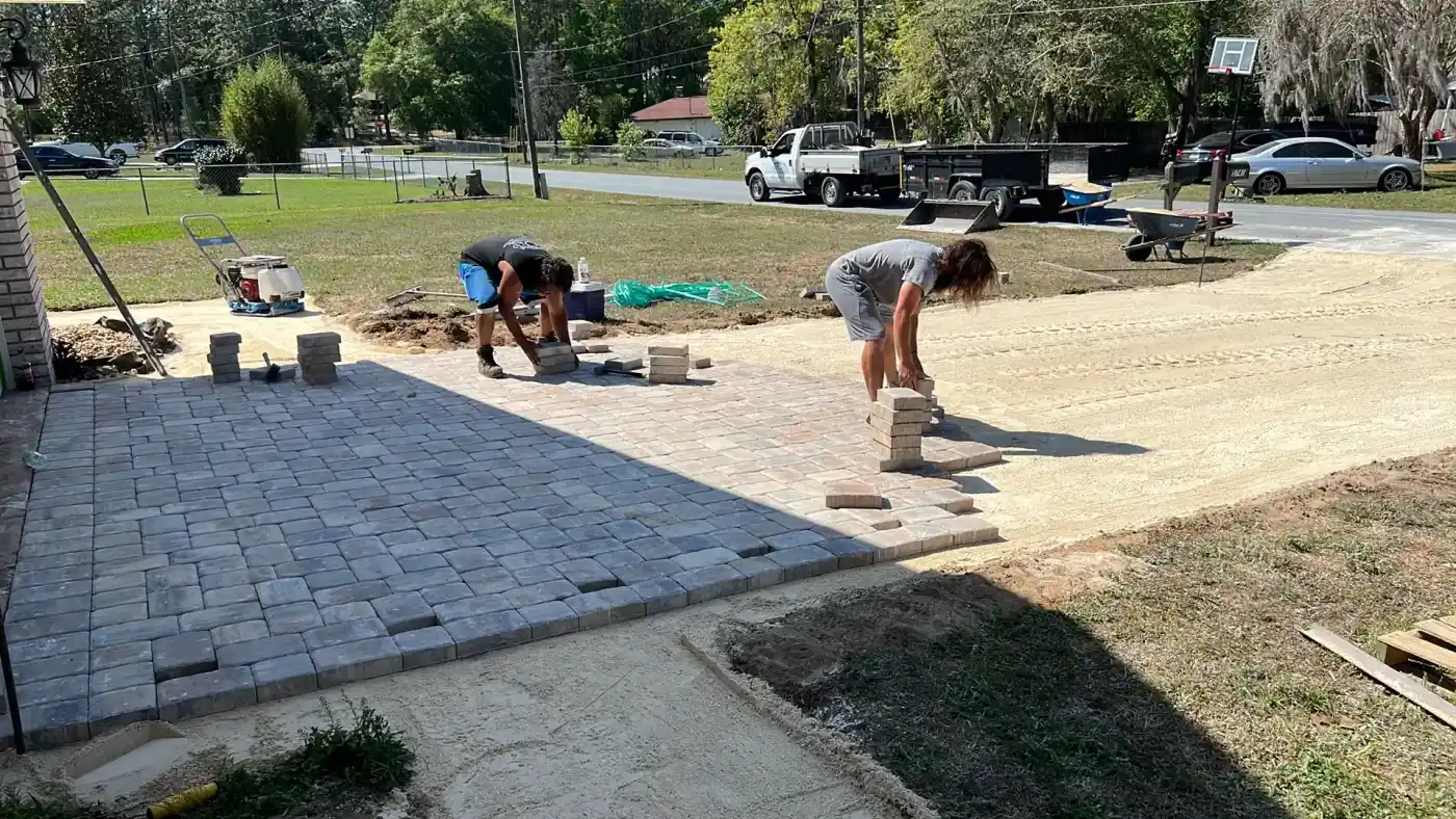 Two people in Hernando County, FL are laying rectangular pavers to construct a patio or driveway on a sunny day, with tools and materials scattered nearby and a truck parked in the background.