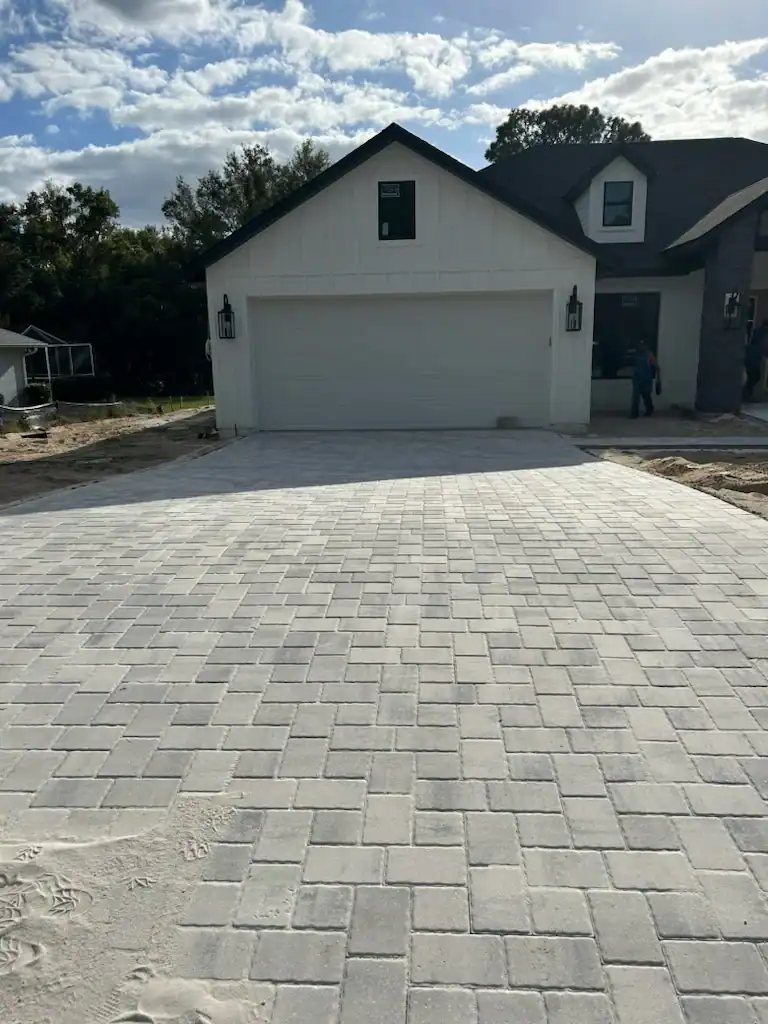 A newly constructed house in Hernando County, FL, features a two-car garage and a wide, light-colored paver driveway under a partly cloudy sky. Landscaper Citrus workers and construction materials are visible near the house.