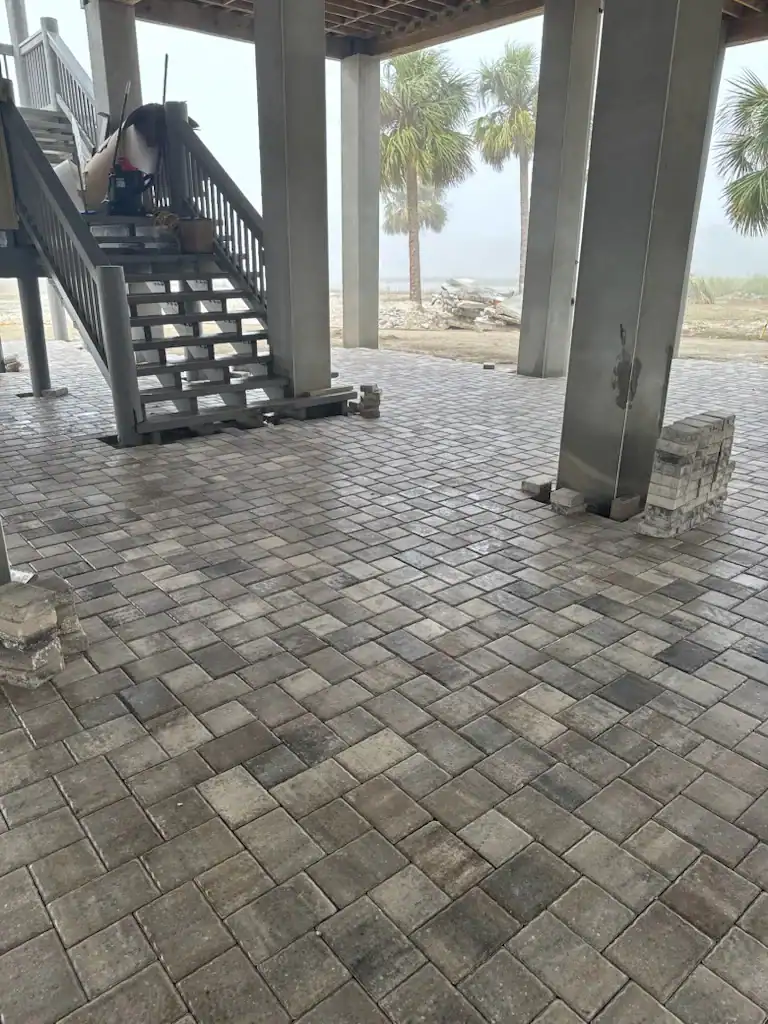 Gray stone pavers laid in a neat, grid-like pattern form a floor beneath a raised structure with stairs and columns. Palm trees and a misty Hernando County, FL landscape are visible in the background. Construction materials are nearby.