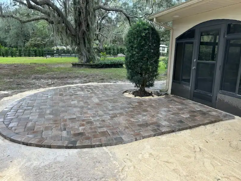 A newly installed curved stone patio by a leading Landscaper Citrus, FL, features a tree planted within a circular cutout. Adjacent to the house with large windows, the patio is bordered by sand, grass, and surrounding trees in Sumter.