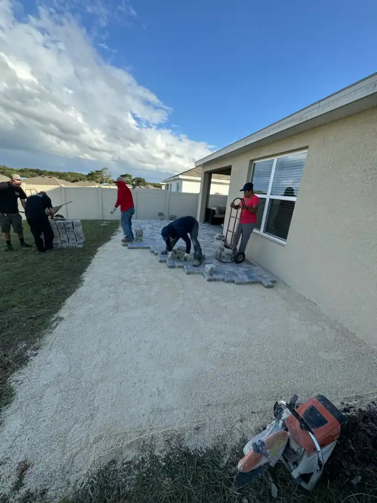 Several people work in a backyard in Hernando County, laying pavers on gravel near a beige house. A Landscaper Citrus crew member uses a dolly to move pavers as others arrange them. A power saw rests on the ground in the foreground.
