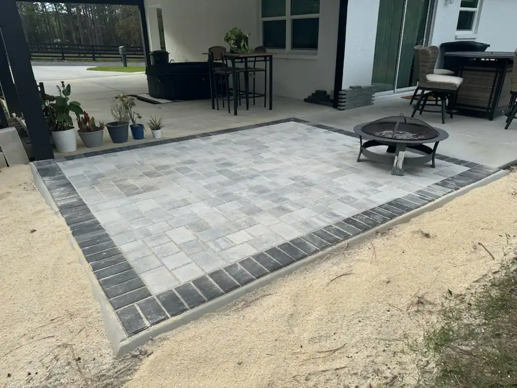A square patio made of gray pavers with a darker border is set in a sandy area next to a house in Hernando County, FL. A metal fire pit sits on the patio, surrounded by several potted plants and outdoor furniture.