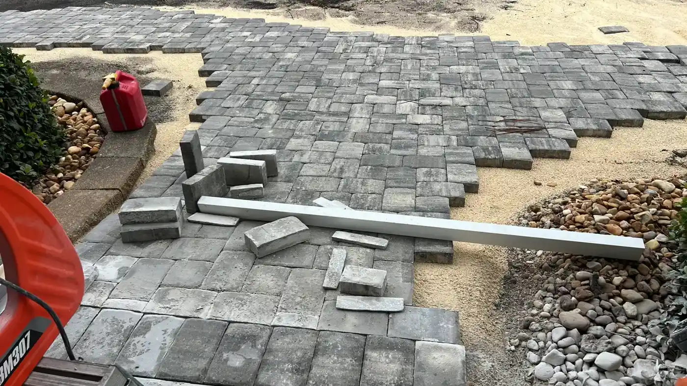 Gray paving stones are partially laid out on a sandy surface in this Landscaper Citrus, FL project. Some stones and a long white plank are scattered on top. Edges remain unfinished, with rocks, exposed dirt, and a red gas can in the upper left.