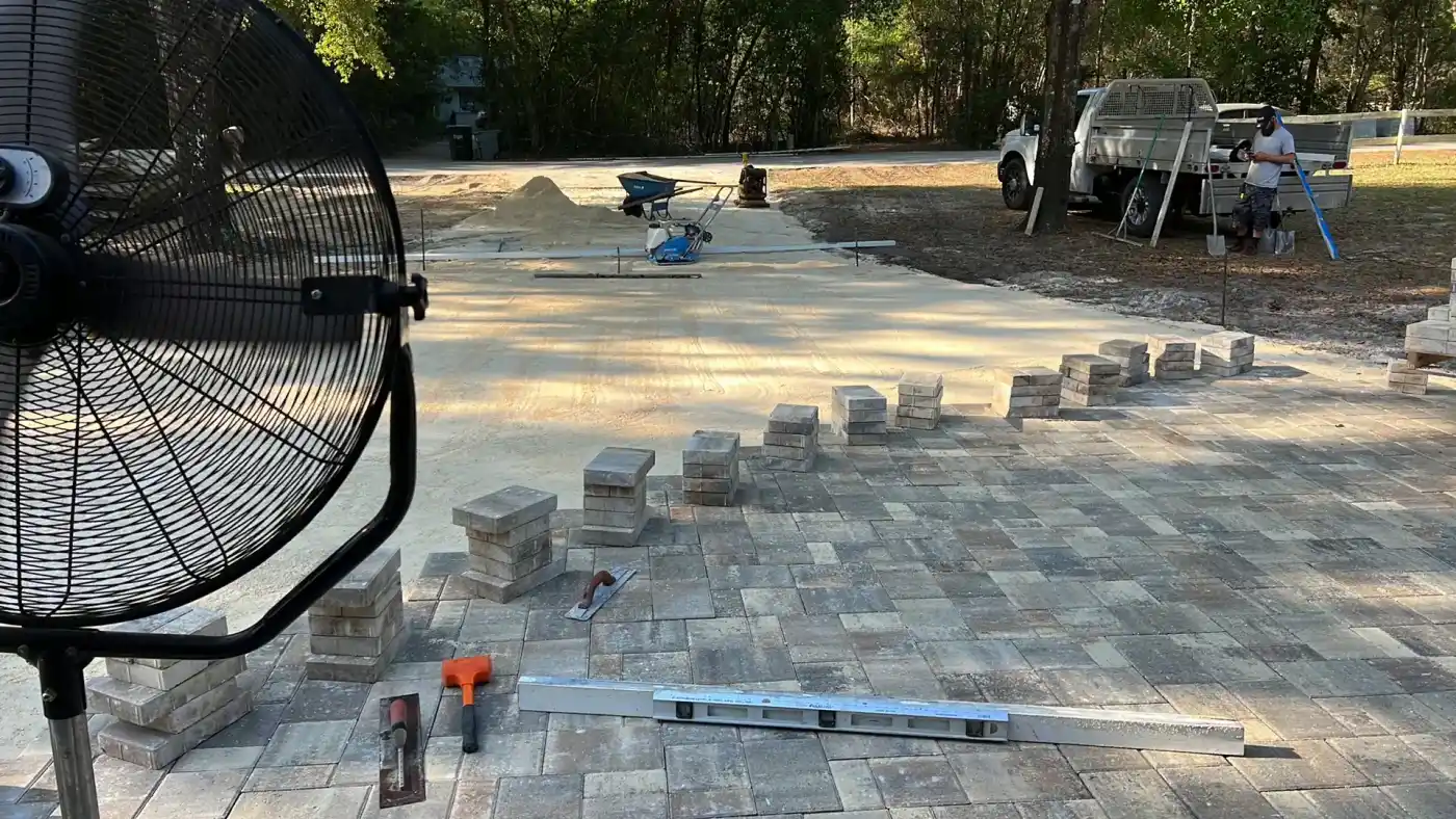 A large fan faces a patio under construction in Hernando County, FL, with paving stones and materials scattered. Stacks of bricks line partly laid pavers, while a Landscaper Citrus truck is parked near the wooded background.
