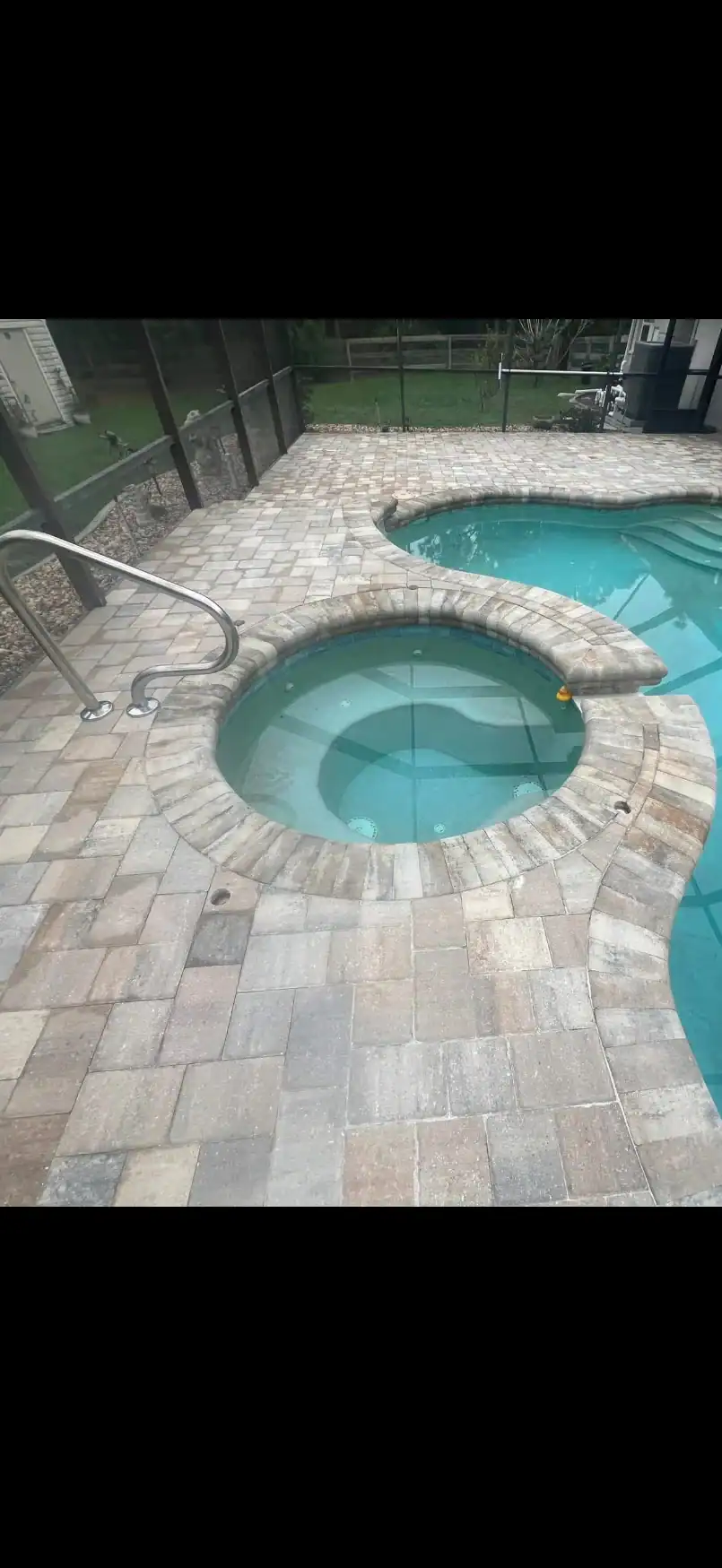 A round hot tub with stone tile surround is connected to a larger swimming pool, both filled with clear water. The pool area, designed by a skilled Landscaper Citrus, FL, is enclosed by a screened structure and bordered by a stone patio.