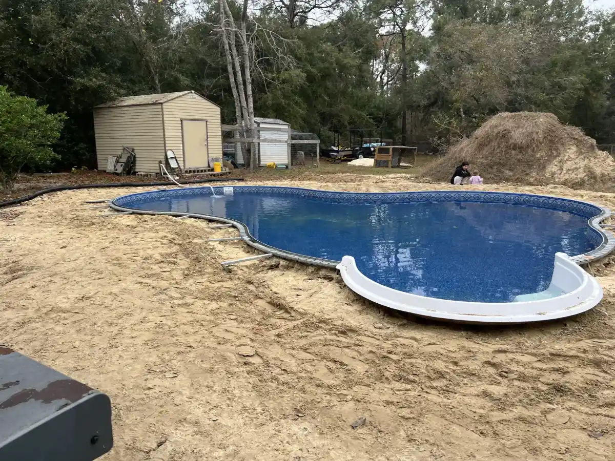 An in-ground pool filled with water sits unfinished in a sandy backyard in Hernando County. There is no surrounding deck, and construction materials are nearby. A shed, piles of dirt, trees, and a person with a child are in the background.
